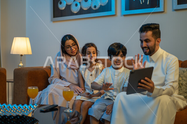 Expressions of happiness and pleasure, watching movies and series using the tablet, making video calls with relatives, a happy family atmosphere at home, a Saudi Gulf Arab couple drinking juice with their children, spending enjoyable family times.