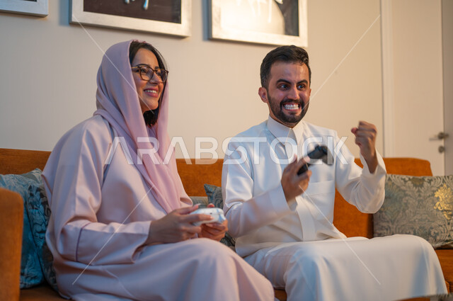 تقنيات الكترونية حديثة ترفيهيه, قضاء وقت ممتع ومسلي في المنزل، التفاعل والاندماج مع ألعاب الفيديو، زوجان عربيان خليجيان سعوديان يمسكان عصا التحكم بتعابير السرور والسعادة، فعاليات عطلة نهاية الأسبوع