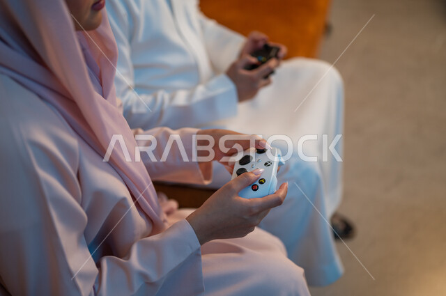 قضاء وقت ممتع ومسلي في المنزل، التفاعل والاندماج مع ألعاب الفيديو، زوجان عربيان خليجيان سعوديان يمسكان عصا التحكم بتعابير السرور والسعادة،  فعاليات عطلة نهاية الأسبوع، تقنيات الكترونية حديثة ترفيهيه