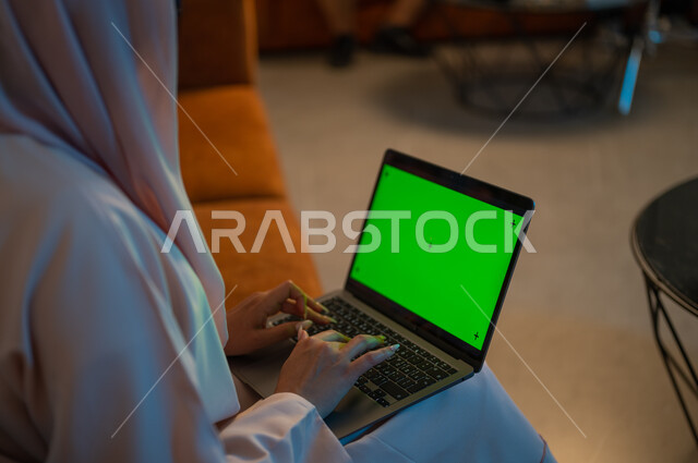 التعلم الإلكتروني من خلال استخدام التقنيات الحديثة، متابعة الأفلام والمسلسلات المفضلة عبر الإنترنت، إجراء مكالمات الفيديو والبثوث المباشرة، صورة مقربة من الخلف لامرأة عربية خليجية سعودية تحمل بيدها جهاز الحاسوب المحمول بشاشة خضراء فارغة،  كروما