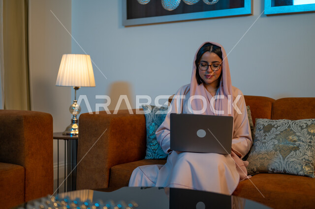 مشاهدة الأفلام والمسلسلات المفضلة عبر الإنترنت، التعلم الإلكتروني من خلال استخدام التقنيات الحديثة، إجراء مكالمات الفيديو والبثوث المباشرة، امرأة عربية خليجية سعودية تجلس على الأريكة في غرفة المعيشة وتحمل جهاز الحاسوب المحمول