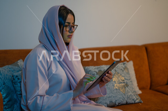 إجراء مكالمات الفيديو والبثوث المباشرة عبر الانترنت, الدردشات والمحادثات مع العائلة والأصدقاء, إنجاز مهام العمل عن بعد, شابة عربية خلجية سعودية تجلس في غرفة المعيشة تحمل بيدها جهاز التابلت, تصفح مواقع النواصل الاجتماعي باستخدام التقنيات الحديثة