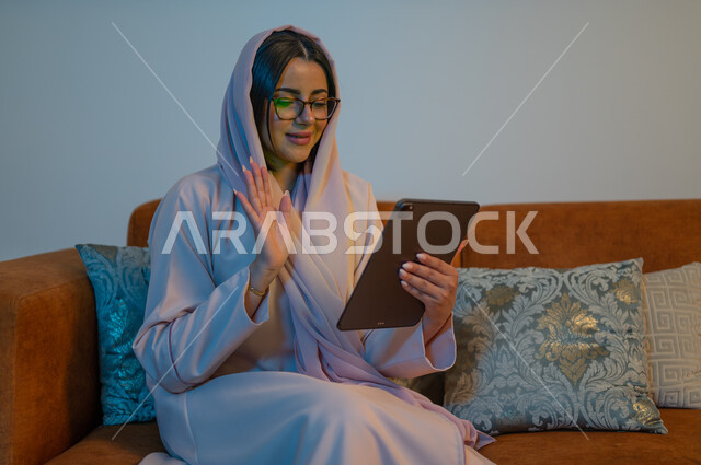 الدردشات والمحادثات مع العائلة والأصدقاء, إجراء مكالمات الفيديو والبثوث المباشرة عبر الانترنت, إنجاز مهام العمل عن بعد, شابة عربية خلجية سعودية تجلس في غرفة المعيشة تحمل بيدها جهاز التابلت, تصفح مواقع النواصل الاجتماعي باستخدام التقنيات الحديثة