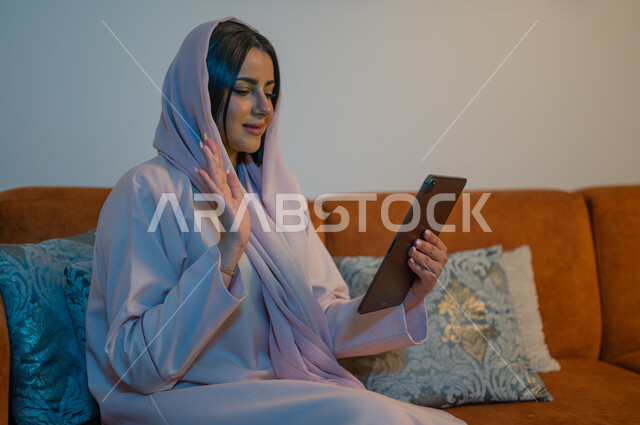 تصفح مواقع النواصل الاجتماعي باستخدام التقنيات الحديثة، الدردشات والمحادثات مع العائلة والأصدقاء, إجراء مكالمات الفيديو والبثوث المباشرة عبر الانترنت, إنجاز مهام العمل عن بعد, شابة عربية خلجية سعودية تجلس في غرفة المعيشة تحمل بيدها جهاز التابلت