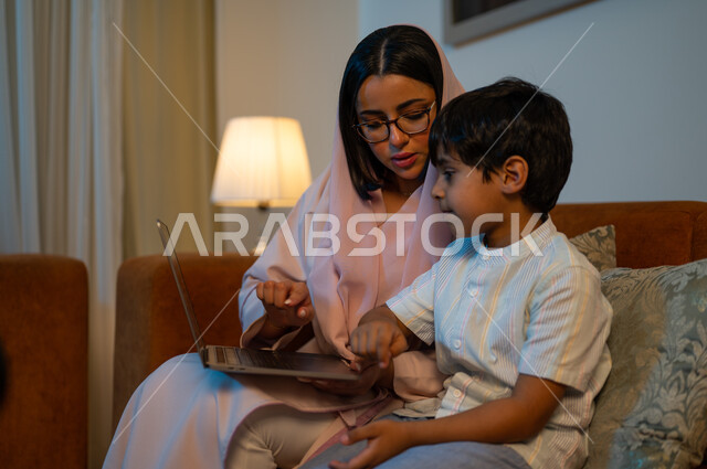 التعليم والدراسة عن بعد، قضاء أوقات ممتعة بمشاهدة الأفلام والمسلسلات المفضلة برفقة العائلة، أم عربية خليجية سعودية تجلس في غرفة المعيشة مع ابنها وتحمل جهاز الحاسوب المحمول، إجراء مكالمات الفيديو والبثوث المباشرة عبر الإنترنت