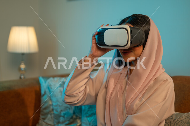 Using modern devices and technologies, gestures to integrate and interact with virtual games, enjoying watching movies of the imaginary world, a Saudi Arabian Gulf woman wearing the hijab and abaya, wearing 3D virtual reality glasses, spending enjoyable times at home.
