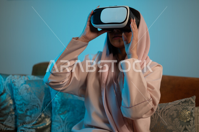 قضاء اوقات ممتعة في المنزل، استخدام الأجهزة والتقنيات الحديثة، إيماءات الاندماج والتفاعل مع الألعاب الافتراضية، الاستمتاع بمشاهدة أفلام العالم الخيالي، إمرأة عربية خليجية سعودية ترتدي الحجاب والعباءة تضع نظارة الواقع الافتراضي ثلاثية الابعاد