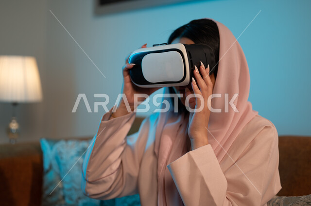الاستمتاع بمشاهدة أفلام العالم الخيالي، قضاء اوقات ممتعة في المنزل، استخدام الأجهزة والتقنيات الحديثة، إيماءات الاندماج والتفاعل مع الألعاب الافتراضية، إمرأة عربية خليجية سعودية ترتدي الحجاب والعباءة تضع نظارة الواقع الافتراضي ثلاثية الابعاد