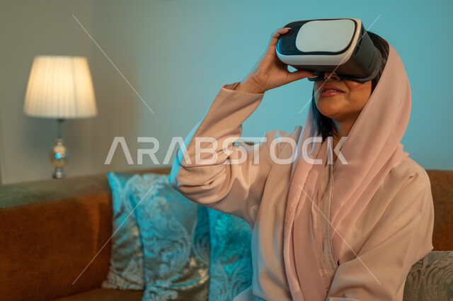 مفهوم التفاعل و الاندماج مع الألعاب الافتراضية، الاستمتاع بمشاهدة أفلام العالم الخيالي، قضاء اوقات ممتعة في المنزل، استخدام الأجهزة والتقنيات الحديثة، إمرأة عربية خليجية سعودية ترتدي الحجاب والعباءة تضع نظارة الواقع الافتراضي ثلاثية الابعاد