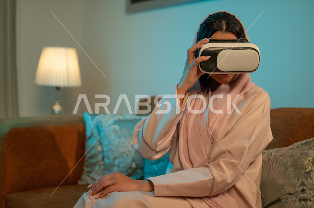 التفاعل و الاندماج مع الألعاب الافتراضية، الاستمتاع بمشاهدة أفلام العالم الخيالي، قضاء اوقات ممتعة في المنزل، استخدام الأجهزة والتقنيات الحديثة، إمرأة عربية خليجية سعودية ترتدي الحجاب والعباءة تضع نظارة الواقع الافتراضي ثلاثية الابعاد