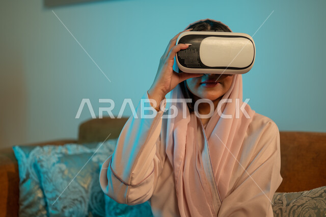 الاستمتاع بمشاهدة أفلام العالم الخيالي، قضاء اوقات ممتعة في المنزل، استخدام الأجهزة والتقنيات الحديثة، إمرأة عربية خليجية سعودية ترتدي الحجاب والعباءة تضع نظارة الواقع الافتراضي ثلاثية الابعاد، التفاعل و الاندماج مع الألعاب الافتراضية