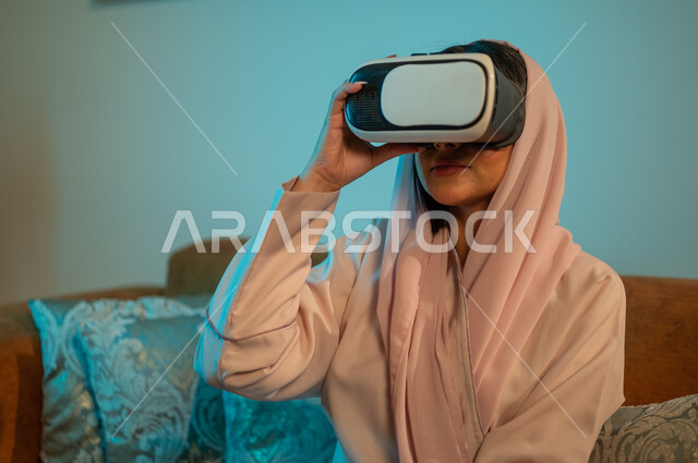 استخدام الأجهزة والتقنيات الحديثة، إمرأة عربية خليجية سعودية ترتدي الحجاب والعباءة تضع نظارة الواقع الافتراضي ثلاثية الابعاد، التفاعل و الاندماج مع الألعاب الافتراضية، الاستمتاع بمشاهدة أفلام العالم الخيالي، قضاء اوقات ممتعة في المنزل