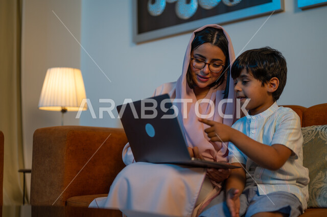 حل الواجبات المنزليه، معلمة خصوصي لمتابعة الدروس في المنزل، أم عربية خليجة سعودية محجبة تقوم بتعليم وتدريس ابنها باستخدام الحاسب المحمول، متابعة منصات التعليم الالكتروني المتاحة عبر الانترنت، دمج التعليم مع التقنية و