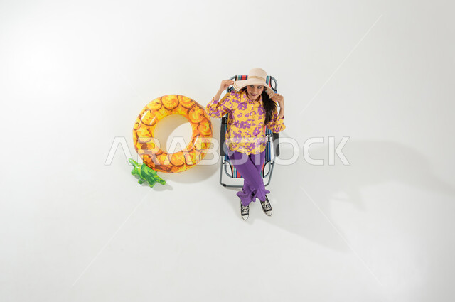 رحلة الشاطئ الممتعة , الاستمتاع بإجازة الصيف , بورتريه علوي عامودي لامرأة عربية خليجية سعودية ترتدي ملابس الشاطئ الصيفية و قبعة من القش تجلس على الكرسي , النظر إلى الكاميرا بإيماءات الفرح و السرور , خلفية بيضاء