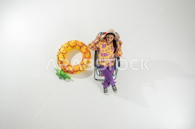 رحلة صيفية ممتعة على الشاطئ , الاستجمام و الاختلاء في الإجازة , بورتريه علوي عامودي لامرأة عربية خليجية سعودية ترتدي ملابس الشاطئ الصيفية و قبعة من القش تجلس على الكرسي , خلفية بيضاء