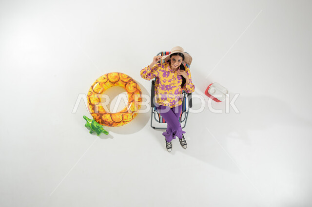 عطلة صيفية ممتعة على الشاطئ , الاستمتاع و الاختلاء في الإجازة , بورتريه علوي عامودي لامرأة عربية خليجية سعودية ترتدي ملابس الشاطئ الصيفية و قبعة من القش تجلس على الكرسي ,إيماءات السعادة و السرور , خلفية بيضاء