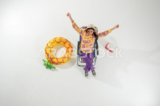 الحماس و السعادة بقدوم فصل الصيف , بورتريه علوي عامودي لامرأة عربية خليجية سعودية ترتدي ملابس الشاطئ الصيفية و قبعة من القش تجلس على الكرسي , قضاء إجازة ممتعة على الشاطئ , خلفية بيضاء