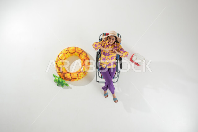 الاستمتاع في وقت الإجازة , أجواء صيفية سعيدة , بورتريه علوي عامودي لامرأة عربية خليجية سعودية ترتدي ملابس الشاطئ الصيفية و قبعة من القش تجلس على الكرسي , النظر إلى الكاميرا بإيماءات الفرح و السرور , خلفية بيضاء