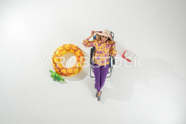 أجواء صيفية سعيدة , بورتريه علوي عامودي لامرأة عربية خليجية سعودية ترتدي ملابس الشاطئ الصيفية و قبعة من القش تجلس على الكرسي , الاستمتاع في وقت الإجازة , النظر إلى الكاميرا بإيماءات الفرح و السرور , خلفية بيضاء