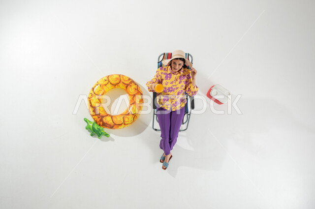 شرب العصائر الباردة و المنعشة , قضاء عطلة الصيف على الشاطئ , بورتريه علوي عامودي لامرأة عربية خليجية سعودية ترتدي ملابس الشاطئ الصيفية و قبعة من القش تجلس على الكرسي , خلفية بيضاء