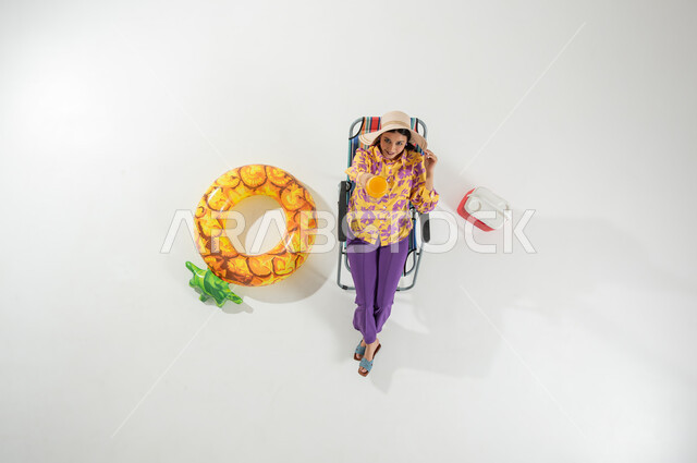 الاستمتاع بالمشروبات الباردة المنعشة , بورتريه علوي عامودي لامرأة عربية خليجية سعودية ترتدي ملابس الشاطئ الصيفية و قبعة من القش تجلس على الكرسي , رحلة صيفية على الشاطئ , خلفية بيضاء