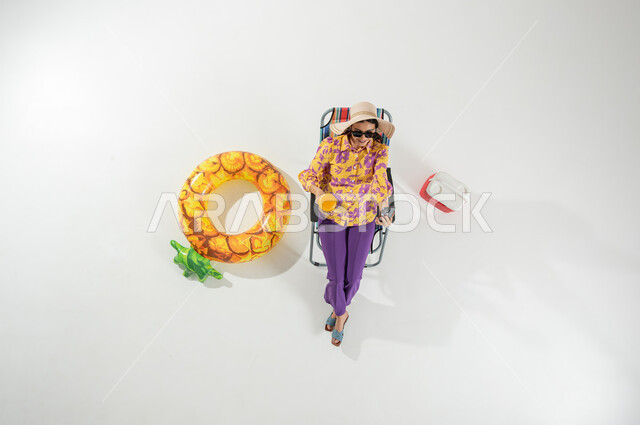 استخدام الهاتف المحمول , قضاء الإجازة على الشاطئ , بورتريه علوي عامودي لامرأة عربية خليجية سعودية ترتدي ملابس الشاطئ الصيفية و قبعة من القش تجلس على الكرسي تمسك هاتفها بيدها , الاستمتاع بعطلة الصيف , خلفية بيضاء