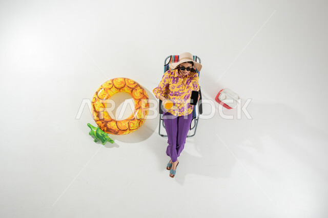 استخدام الأجهزة الحديثة , الاستمتاع بعطلة الصيف , بورتريه علوي عامودي لامرأة عربية خليجية سعودية ترتدي ملابس الشاطئ الصيفية و قبعة من القش تجلس على الكرسي تمسك هاتفها بيدها , عرض شاشة سوداء فارغة عبر الجوال  , خلفية بيضاء