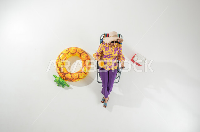 التواصل مع الأصحاب عبر الهاتف المحمول , بورتريه علوي عامودي لامرأة عربية خليجية سعودية ترتدي ملابس الشاطئ الصيفية و قبعة من القش تجلس على الكرسي تمسك هاتفها بيدها , قضاء الإجازة على الشاطئ , خلفية بيضاء