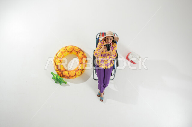 عطلة صيفية ممتعة , التقاط الصور التذكارية , بورتريه علوي عامودي لامرأة عربية خليجية سعودية ترتدي ملابس الشاطئ الصيفية و قبعة من القش تجلس على الكرسي تمسك كاميرا التصوير بيديها , الاستمتاع برحلة الشاطئ , خلفية بيضاء