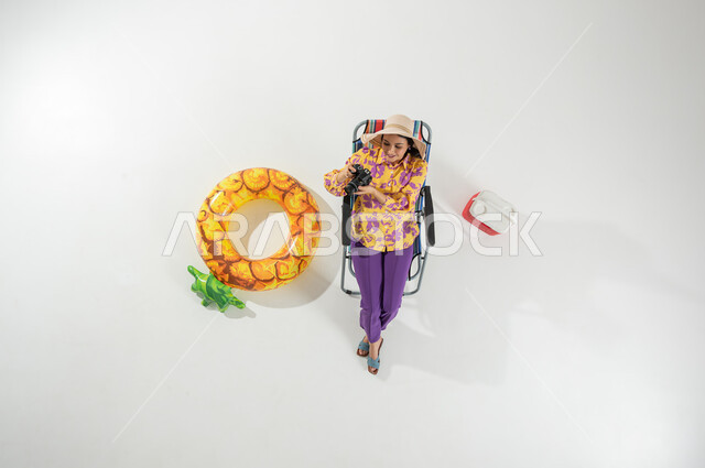 الاستمتاع برحلة الشاطئ , التقاط الصور التذكارية , بورتريه علوي عامودي لامرأة عربية خليجية سعودية ترتدي ملابس الشاطئ الصيفية و قبعة من القش تجلس على الكرسي تمسك كاميرا التصوير بيديها , , عطلة صيفية ممتعة , خلفية بيضاء