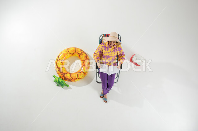 Practicing the hobby of reading during the summer vacation, a vertical portrait of a Saudi Gulf Arab woman wearing summer beach clothes and a straw hat, sitting on a chair holding a book in her hands, enjoying a beach trip, white background