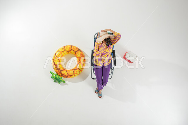 الاستمتاع بمشاهدة منظر البحر  , رؤية الأشياء البعيدة عبر المنظار  , بورتريه علوي عامودي لامرأة عربية خليجية سعودية ترتدي ملابس الشاطئ الصيفية و قبعة من القش تجلس على الكرسي تمسك بيديها منظار ثنائي العينين , خلفية بيضاء