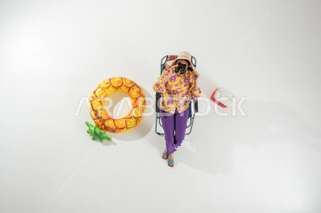 رؤية الأشياء البعيدة عبر المنظار , الاستمتاع بمشاهدة منظر البحر , بورتريه علوي عامودي لامرأة عربية خليجية سعودية ترتدي ملابس الشاطئ الصيفية و قبعة من القش تجلس على الكرسي تمسك بيديها منظار ثنائي العينين , خلفية بيضاء