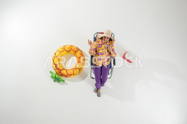 تنمية المواهب في الإجازة , قضاء عطلة الصيف على الشاطئ , بورتريه علوي عامودي لامرأة عربية خليجية سعودية ترتدي ملابس الشاطئ الصيفية و قبعة من القش تجلس على الكرسي تمسك المايكروفون بيدها , خلفية بيضاء