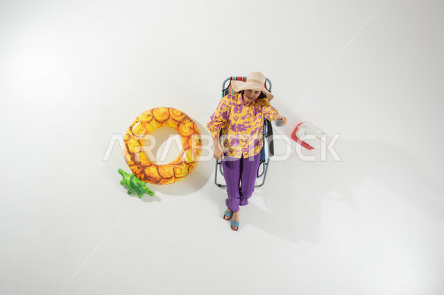 ممارسة هواية الغناء في الإجازة , الاستمتاع برحلات الشاطئ , بورتريه علوي عامودي لامرأة عربية خليجية سعودية ترتدي ملابس الشاطئ الصيفية و قبعة من القش تجلس على الكرسي تمسك المايكروفون بيدها , خلفية بيضاء