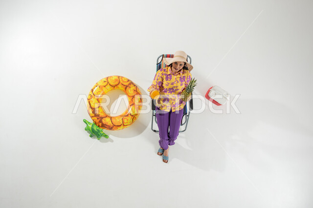 الاسترخاء و الاستمتاع بالعطلة الصيفية , بورتريه علوي عامودي لامرأة عربية خليجية سعودية ترتدي ملابس الشاطئ الصيفية و قبعة من القش تجلس على الكرسي و تمسك فاكهة الأناناس و كأس العصير بيديها , رحلة الشاطئ الممتعة , خلفية بيضاء