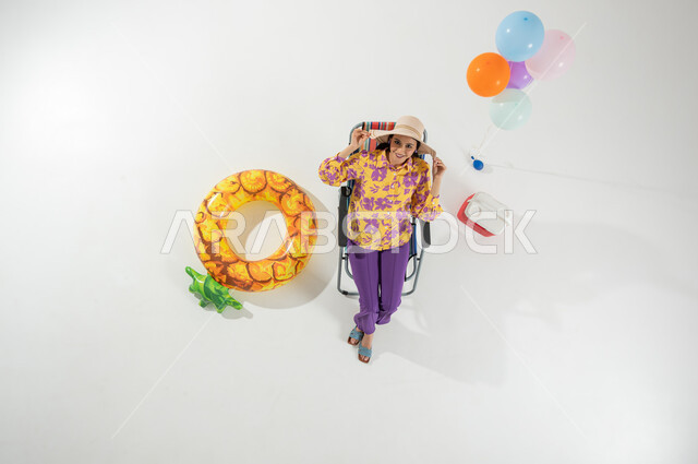 الاستمتاع بالاستجمام على الشاطئ , بورتريه علوي عامودي لامرأة عربية خليجية سعودية ترتدي ملابس الشاطئ الصيفية و قبعة من القش , قضاء الإجازة الصيفية بالرحلات الممتعة , خلفية بيضاء