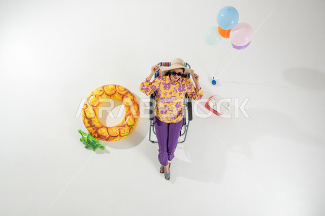 الاستجمام و الاختلاء في العطلة , بورتريه علوي عامودي لامرأة عربية خليجية سعودية ترتدي ملابس الشاطئ الصيفية و نظارة شمسية تمسك قبعة من القش بيديها  , إجازة صيفية على الشاطئ , خلفية بيضاء