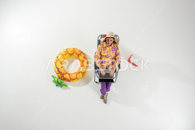 استخدام التكنولوجيا في الترفيه أثناء العطلة , إجازة صيفية على الشاطئ , بورتريه علوي عامودي لامرأة عربية خليجية سعودية ترتدي ملابس الشاطئ الصيفية و قبعة شمسية تقوم بالكتابة على لوحة مفاتيح جهاز الحاسوب , خلفية بيضاء