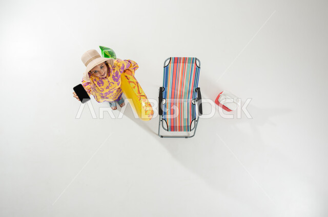 استخدام الهاتف المحمول في الإجازة للتسلية , بورتريه علوي عامودي لامرأة عربية خليجية سعودية ترتدي ملابس الشاطئ الصيفية و قبعة شمسية تحمل عوامة السباحة و الجوال بيديها , الاستمتاع بالإجازة على الشاطئ , خلفية بيضاء