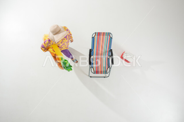 التسلية و الترفيه في رحلات الشاطئ, بورتريه علوي عامودي لامرأة عربية خليجية سعودية ترتدي ملابس الشاطئ الصيفية و قبعة شمسية تحمل عوامة السباحة بيدها , الاستمتاع بوقت العطلة, خلفية بيضاء