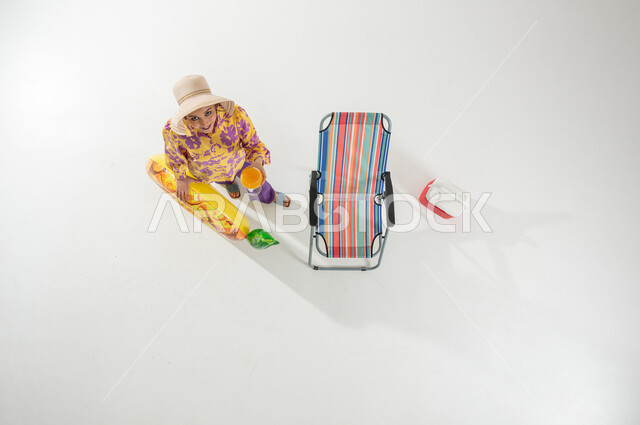 الاستمتاع بالمشروبات الباردة و المنعشة , بورتريه علوي عامودي لامرأة عربية خليجية سعودية ترتدي ملابس الشاطئ الصيفية و قبعة شمسية تحمل عوامة السباحة و كأس العصير بيديها , النظر إلى الكاميرا بإيماءات الفرح و السرور , خلفية بيضاء