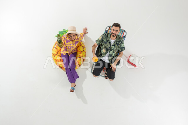 رحلات الشاطئ الممتعة , الاستمتاع بالمشروبات و الفواكه الصيفية  , بورتريه علوي عامودي لزوجان عربيان خليجيان سعوديان يرتديان ملابس الشاطئ الصيفية , النظر إلى الكاميرا بإيماءات الفرح و السرور , خلفية بيضاء