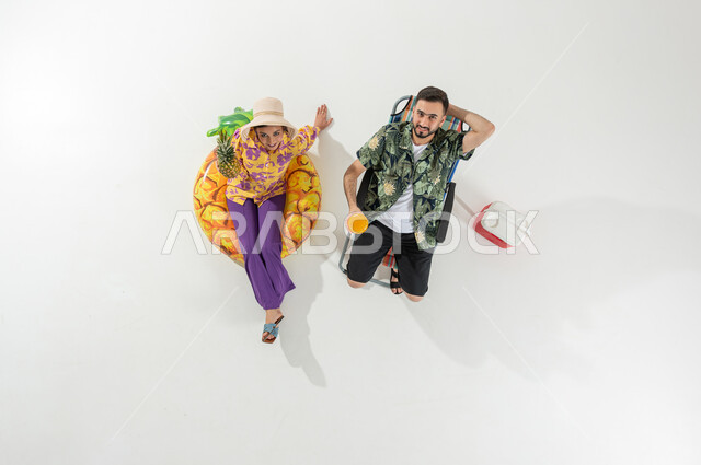 A tourist trip on the beach, enjoying summer drinks and fruits, a vertical overhead portrait of a Saudi Gulf Arab couple wearing summer beach clothes, spending fun times on vacation, white background