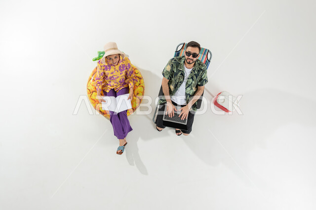 ممارسة الهوايات في إجازة الصيف , استخدام الأجهزة التقنية للترفيه , بورتريه علوي عامودي لزوجان عربيان خليجيان سعوديان يرتديان ملابس الشاطئ الصيفية , الاستمتاع بالعطلة على الشاطئ , خلفية بيضاء