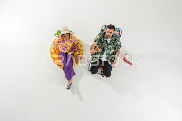 الاستمتاع بالفواكه الصيفية , قضاء وقت الإجازة على الشاطئ , بورتريه علوي عامودي لزوجان عربيان خليجيان سعوديان يرتديان ملابس الشاطئ الصيفية يأكلان فاكهة البطيخ , النظر إلى الكاميرا بإيماءات الفرح و السرور , خلفية بيضاء