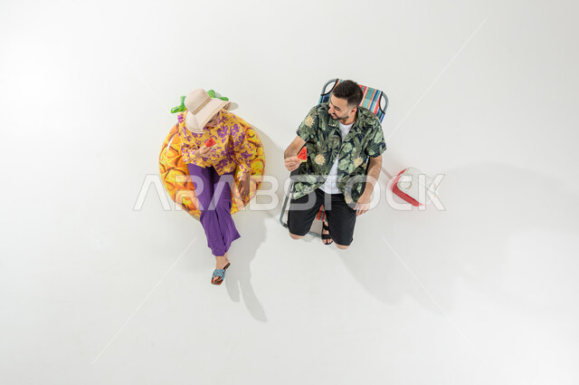 فعاليات للتسلية و الترفيه , مفهوم المحبة و الالفة بين الزوجين , بورتريه علوي عامودي لزوجان عربيان خليجيان سعوديان يرتديان ملابس الشاطئ الصيفية يأكلان فاكهة البطيخ , قضاء وقت العطلة على الشاطئ, خلفية بيضاء