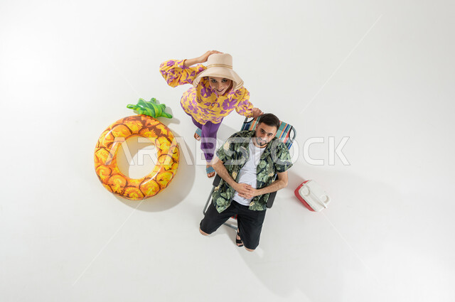 قضاء وقت العطلة على الشاطئ , فعاليات للترفيه و التسلية , بورتريه علوي عامودي لزوجان عربيان خليجيان سعوديان يرتديان ملابس الشاطئ الصيفية , النظر إلى الكاميرا بإيماءات الفرح و السرور , خلفية بيضاء