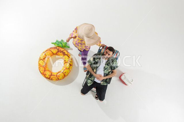 The concept of affection and intimacy between spouses, summer vacation on the beach, a vertical overhead portrait of a Saudi Gulf Arab couple wearing summer beach clothes, a man sitting on a chair looking at his wife with loving gestures, white background