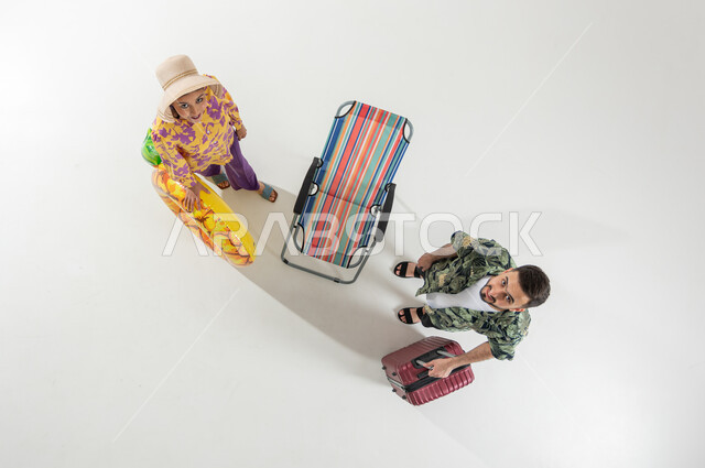 قضاء أوقات الإجازة بالترفيه و الاستمتاع , بورتريه علوي عامودي لزوجان عربيان خليجيان سعوديان يرتديان ملابس الشاطئ الصيفية يحملان حقيبة سفر و عوامة السباحة , الرحلات و السفر في السعودية, خلفية بيضاء